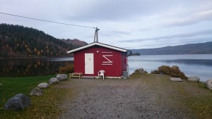 Liten rød bygning med Holsfjorden i bakgrunn