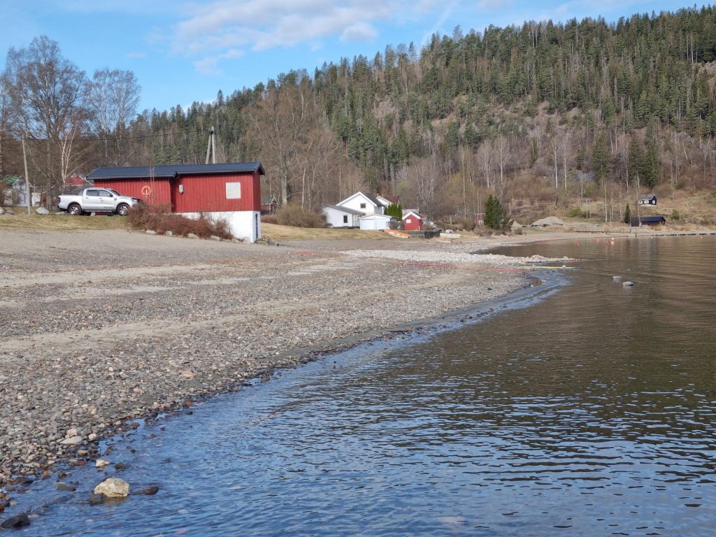 Bilde: Et lite rødt hus, strand og vann. Stranden er høyrer enn normalt